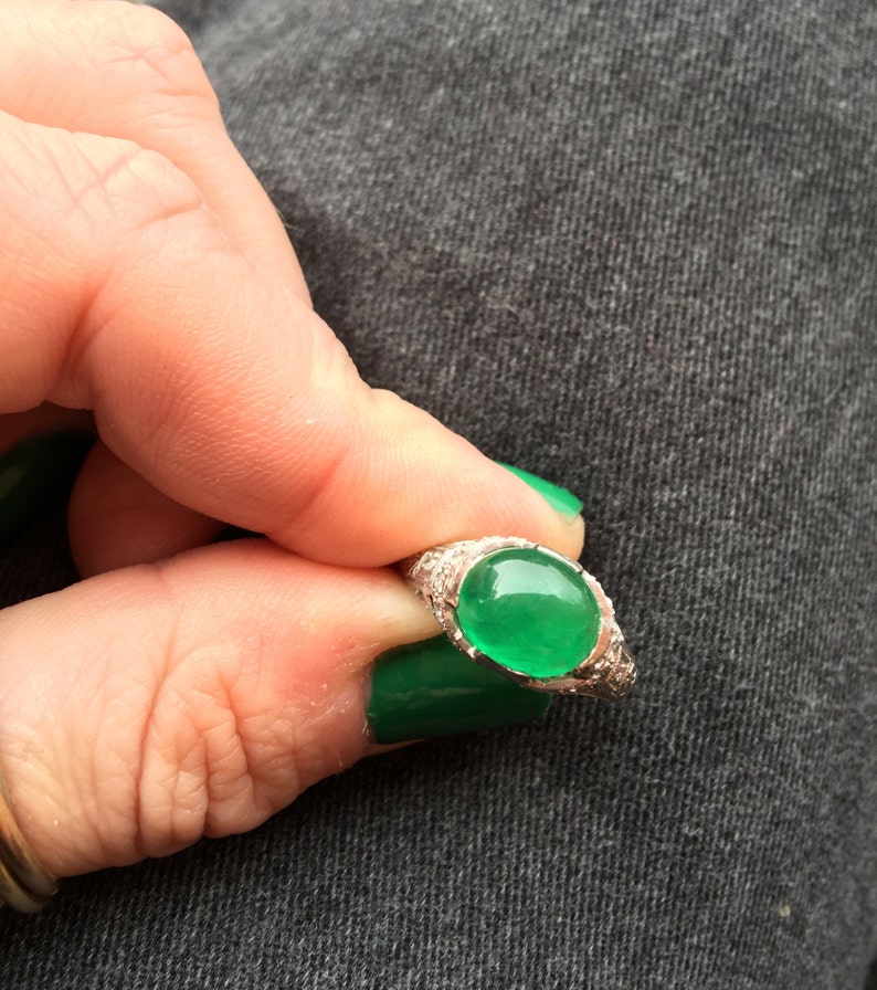 May include: A silver ring with a green oval gemstone set in a bezel setting. The ring is surrounded by small, clear gemstones.