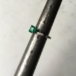 May include: A silver ring with a square emerald green gemstone and small diamonds on the band. The ring is sized on a ring sizer that shows a size 6.