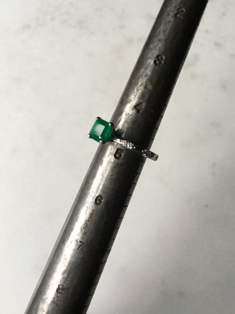 May include: A silver ring with a square emerald green gemstone and small diamonds on the band. The ring is sized on a ring sizer that shows a size 6.