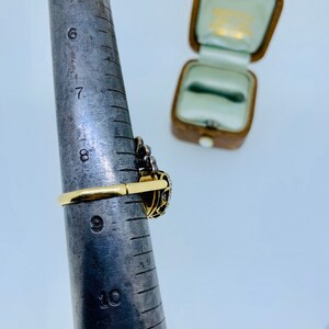 May include: A gold ring with a dark gemstone and intricate filigree design is shown on a ring sizer. The ring is set with small, clear stones. An open, vintage-style ring box is in the background.