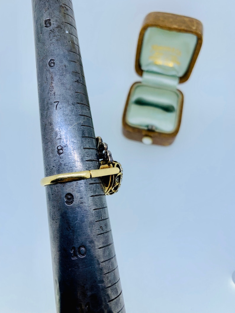 May include: A gold ring with a dark gemstone and intricate filigree design is shown on a ring sizer. The ring is set with small, clear stones. An open, vintage-style ring box is in the background.