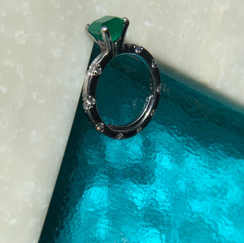 May include: A silver ring with a large, emerald green gemstone and smaller, clear gemstones around the band.