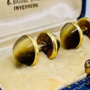 K&ouml;nnte beinhalten: Ein Paar goldene Manschettenkn&ouml;pfe mit braunen und goldenen Tigeraugen-Steinen. Die Manschettenkn&ouml;pfe befinden sich in einer blauen und wei&szlig;en Schachtel.