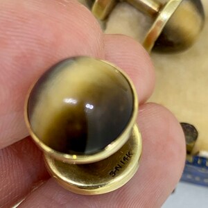 K&ouml;nnte beinhalten: Ein Paar goldene Manschettenkn&ouml;pfe mit braunen und wei&szlig;en Steinen. Die Manschettenkn&ouml;pfe sind mit "3N114K" gekennzeichnet.