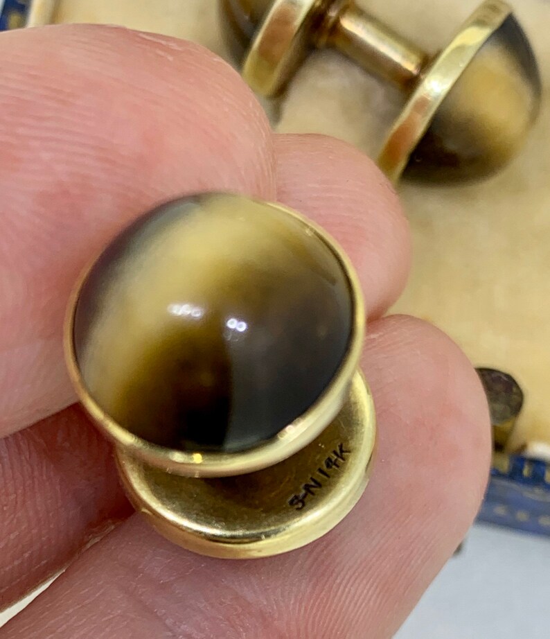 K&ouml;nnte beinhalten: Ein Paar goldene Manschettenkn&ouml;pfe mit braunen und wei&szlig;en Steinen. Die Manschettenkn&ouml;pfe sind mit "3N114K" gekennzeichnet.