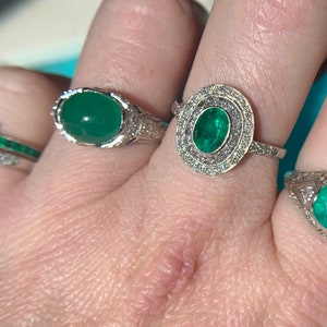 May include: Three silver rings with emerald gemstones. The rings are on a person's finger. The center ring has a large emerald surrounded by smaller diamonds.