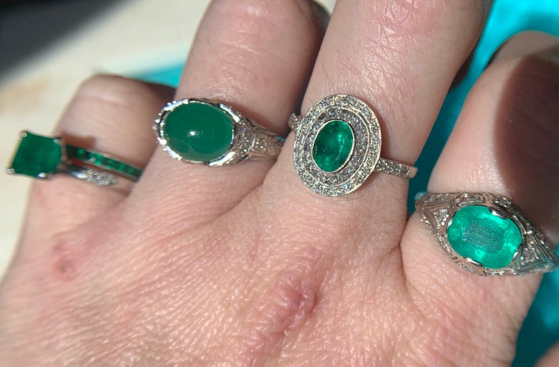 May include: Three silver rings with emerald gemstones. The rings are on a person's finger. The center ring has a large emerald surrounded by smaller diamonds.