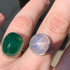 May include: Two rings with gemstones. The ring on the left has a green oval-shaped gemstone set in a gold band. The ring on the right has a light blue star-shaped gemstone set in a silver band.