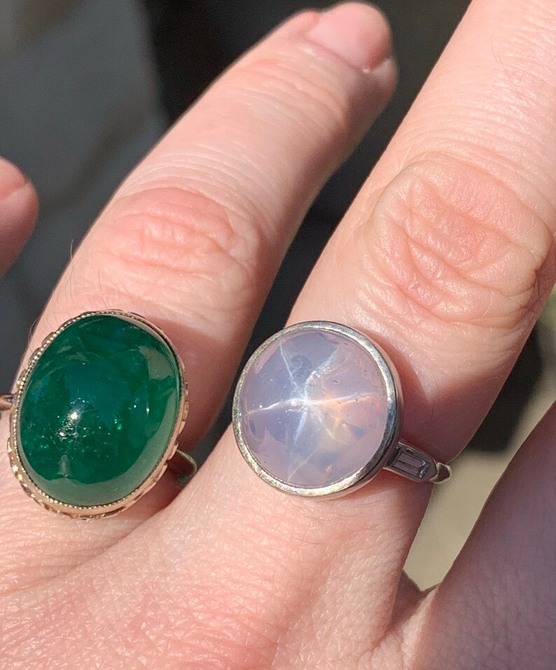 May include: Two rings with gemstones. The ring on the left has a green oval-shaped gemstone set in a gold band. The ring on the right has a light blue star-shaped gemstone set in a silver band.