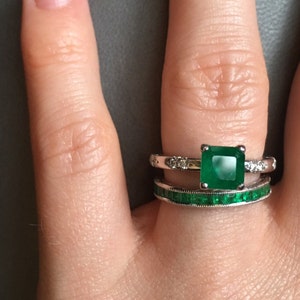 May include: A close-up of a hand wearing two rings. The top ring features a large, square emerald gemstone surrounded by smaller diamonds. The bottom ring is a simple band with emerald gemstones.
