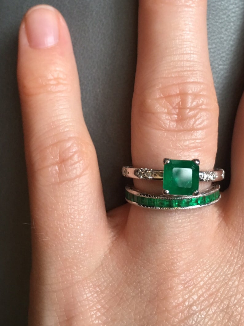 May include: A close-up of a hand wearing two rings. The top ring features a large, square emerald gemstone surrounded by smaller diamonds. The bottom ring is a simple band with emerald gemstones.