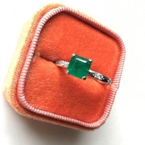 May include: A ring with a large emerald green gemstone surrounded by smaller diamonds set in a white metal band. The ring is displayed in a small orange velvet ring box.