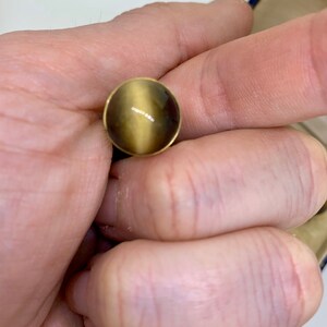 K&ouml;nnte beinhalten: Ein runder, goldfarbener Ring mit einem braunen und goldenen Edelstein. Der Edelstein hat einen Chatoyance-Effekt, d. h. er scheint je nach Lichteinfall die Farbe zu &auml;ndern.