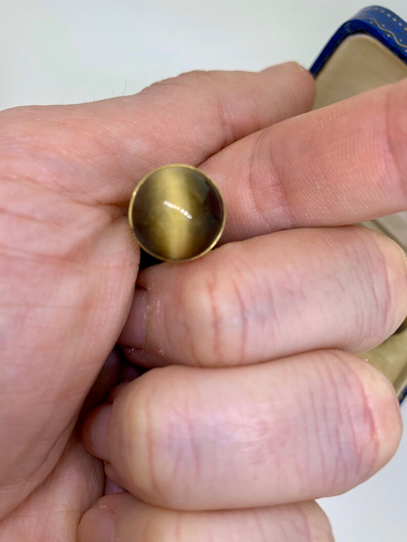 K&ouml;nnte beinhalten: Ein runder, goldfarbener Ring mit einem braunen und goldenen Edelstein. Der Edelstein hat einen Chatoyance-Effekt, d. h. er scheint je nach Lichteinfall die Farbe zu &auml;ndern.