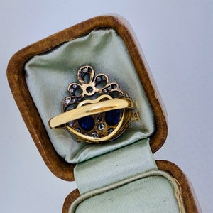 May include: A gold ring with two oval blue gemstones and small diamonds, set in a heart-shaped design. The ring is displayed in a vintage, brown, hinged jewelry box with a light green satin lining.