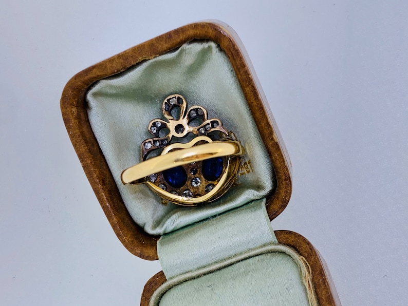 May include: A gold ring with two oval blue gemstones and small diamonds, set in a heart-shaped design. The ring is displayed in a vintage, brown, hinged jewelry box with a light green satin lining.