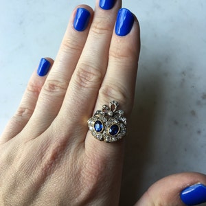 May include: A gold-toned ring with two oval blue gemstones and numerous smaller clear stones. The ring is on a hand with bright blue nail polish.