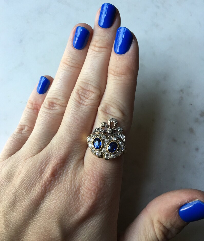 May include: A gold-toned ring with two oval blue gemstones and numerous smaller clear stones. The ring is on a hand with bright blue nail polish.