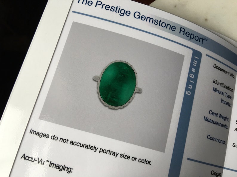 May include: A green gemstone ring with a silver setting. The ring is oval shaped and has a detailed design around the edge of the stone. The ring is photographed on a white background.