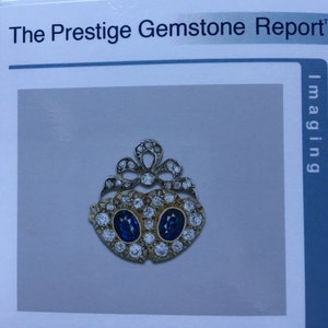 May include: A gold-toned gemstone pendant with two oval, deep blue gemstones at the center, surrounded by smaller, clear gemstones. The pendant has a decorative, openwork design at the top. The text "The Prestige Gemstone Report" is visible.