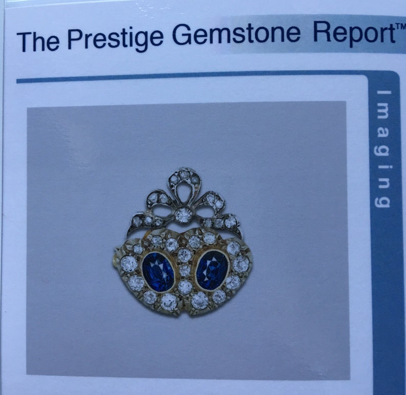 May include: A gold-toned gemstone pendant with two oval, deep blue gemstones at the center, surrounded by smaller, clear gemstones. The pendant has a decorative, openwork design at the top. The text "The Prestige Gemstone Report" is visible.