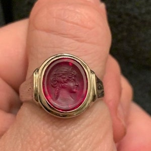 Puede incluir: Un anillo de plata con una piedra preciosa roja de forma ovalada que presenta un perfil tallado de la cara de una mujer.
