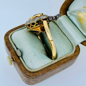 May include: A gold ring with a central dark blue gemstone, surrounded by smaller clear stones. The ring is in a vintage brown jewelry box with a light blue interior. The ring's band is a simple, polished gold.