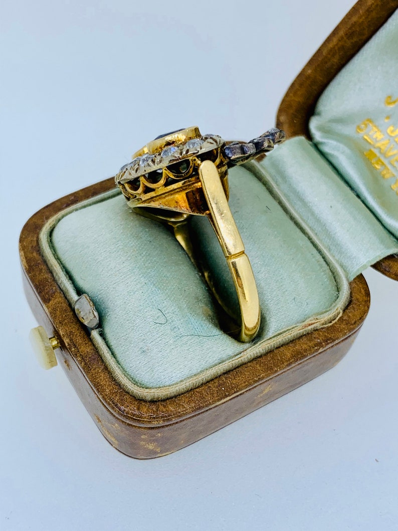 May include: A gold ring with a central dark blue gemstone, surrounded by smaller clear stones. The ring is in a vintage brown jewelry box with a light blue interior. The ring's band is a simple, polished gold.