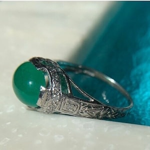 May include: A silver ring with a green gemstone and intricate filigree detailing. The ring is set on a blue background.