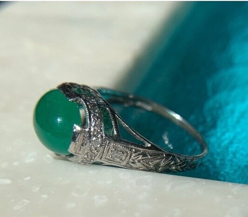 May include: A silver ring with a green gemstone and intricate filigree detailing. The ring is set on a blue background.