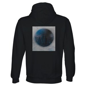Puede incluir: Una sudadera con capucha negra que presenta un gráfico cuadrado en la parte posterior. El gráfico muestra un diseño circular en degradado azul y negro con tres siluetas indistintas de figuras sobre un fondo gris claro texturizado.