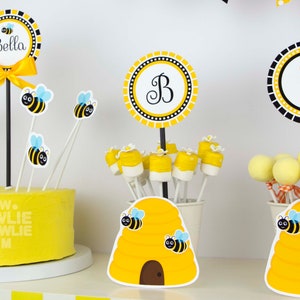May include: A yellow and black bee-themed birthday party centerpiece with a cake, beehive-shaped containers, and cake pops. The cake is decorated with bee-themed toppers and the text "www.owliepowlie.com". The containers are decorated with beehive designs and bee stickers. The cake pops are decorated with yellow and black bee designs. The centerpiece also includes a yellow and black circle with the letter "B" and a yellow and black circle with the number "1".