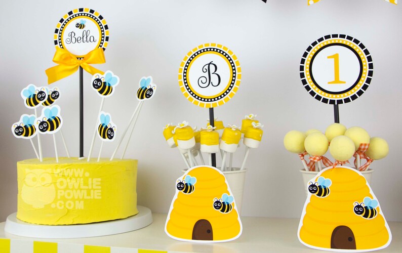 May include: A yellow and black bee-themed birthday party centerpiece with a cake, beehive-shaped containers, and cake pops. The cake is decorated with bee-themed toppers and the text "www.owliepowlie.com". The containers are decorated with beehive designs and bee stickers. The cake pops are decorated with yellow and black bee designs. The centerpiece also includes a yellow and black circle with the letter "B" and a yellow and black circle with the number "1".
