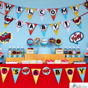 May include: A superhero-themed party display featuring two rows of triangular pennant banners with comic book style graphics and the text WELCOME BABY and IT'S A BOY. The table includes water bottles, food labels, and decorative cutouts.