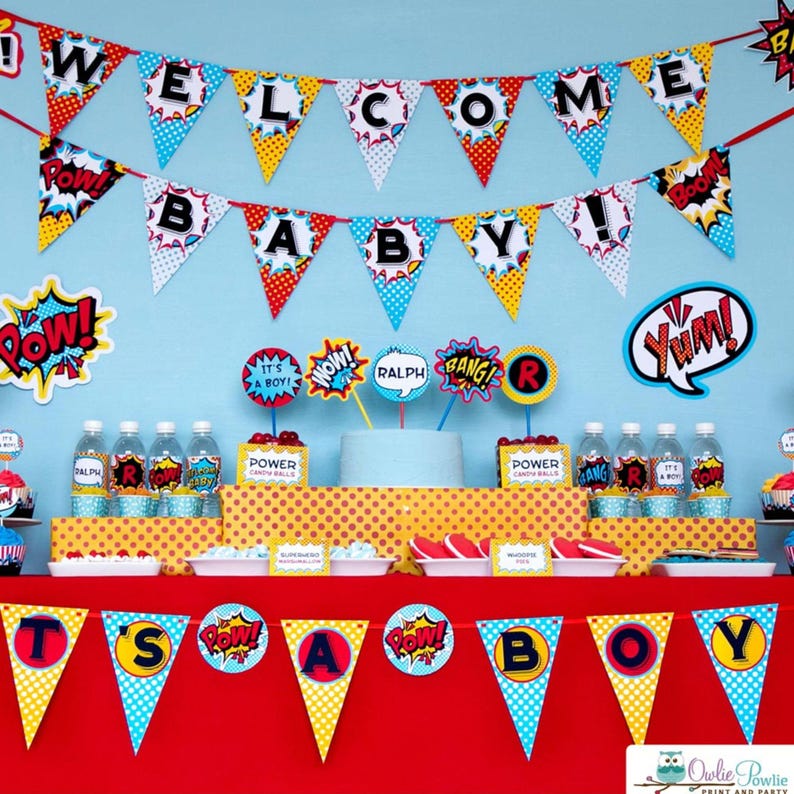 May include: A superhero-themed party display featuring two rows of triangular pennant banners with comic book style graphics and the text WELCOME BABY and IT'S A BOY. The table includes water bottles, food labels, and decorative cutouts.