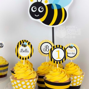 May include: Yellow and black striped cupcake wrappers with white polka dots. Cupcakes are topped with yellow frosting and bee-themed toppers. Toppers include a bee, the letter "B", the number "1", and the text "Happy Bee-day".