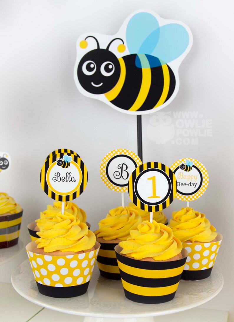 May include: Yellow and black striped cupcake wrappers with white polka dots. Cupcakes are topped with yellow frosting and bee-themed toppers. Toppers include a bee, the letter "B", the number "1", and the text "Happy Bee-day".
