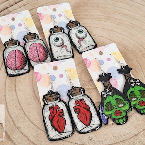 May include: Four pairs of Halloween-themed earrings. The first pair features a brain in a jar, the second pair features an eyeball in a jar, the third pair features a heart in a jar, and the fourth pair features a green zombie head.