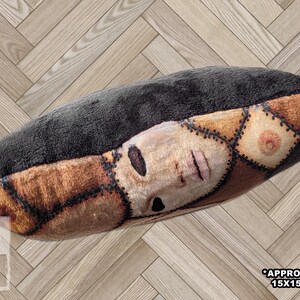 Side view of handmade square pillow on wood floor made of minky fleece fabric featuring an original patchwork print of multiple shades of human skin and body parts stitched together including haunted faces, ears, elbows, and nipples. Solid black back