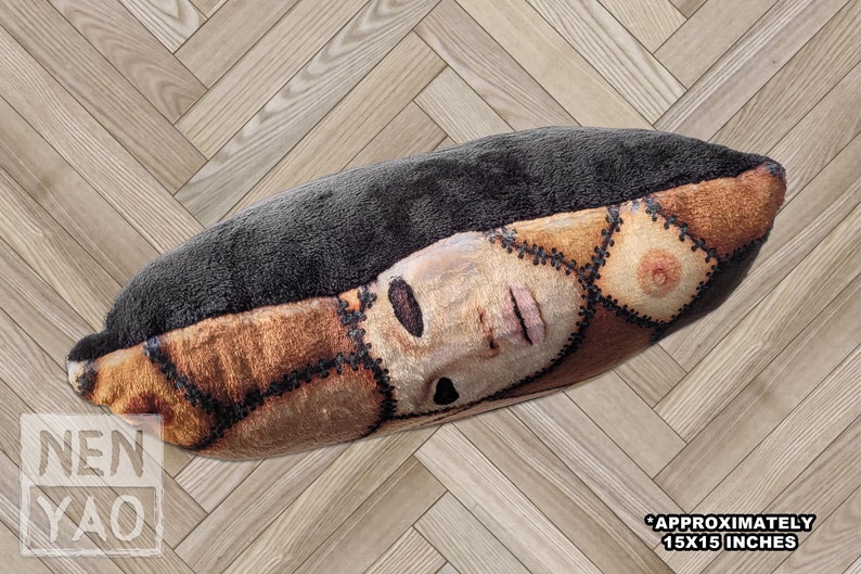 Side view of handmade square pillow on wood floor made of minky fleece fabric featuring an original patchwork print of multiple shades of human skin and body parts stitched together including haunted faces, ears, elbows, and nipples. Solid black back