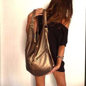 Pode incluir: Uma grande bolsa hobo de couro de cor dourada com um bolso com zíper. A bolsa é usada no ombro de uma pessoa com cabelo castanho longo.