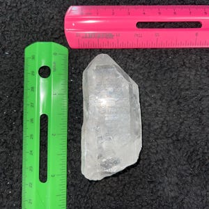 May include: A clear quartz crystal point with a rough, natural texture. The crystal is approximately 2 inches long and is resting on a black surface.