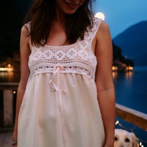 Può includere: Una donna indossa un abito bianco senza maniche in pizzo all'uncinetto con un fiocco rosa in vita.
