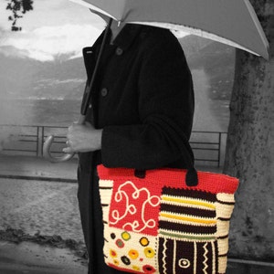 May include: A woman is walking in the rain with a black coat and a colorful crocheted tote bag. The bag has a red, white, black, and yellow patchwork design.
