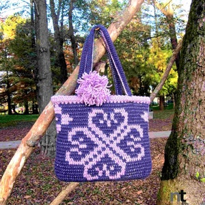 Può includere: Una borsa a mano in crochet viola e rosa con un designo floreale. La borsa ha un pon pon rosa sul manico.