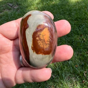 May include: A smooth, polished, oval-shaped stone featuring a multi-colored surface with a large, prominent dark reddish-brown patch centered on a light grey background, with a smaller, vibrant orange-yellow textured area within the brown section.