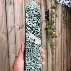 May include: A tall, six-sided crystal tower with a pointed top. The stone features a mottled pattern of deep forest green and white mineral inclusions, creating a dense, speckled appearance across the entire vertical surface of the polished object.