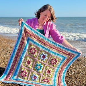 Op de afbeelding: Een vrouw houdt een kleurrijk gehaakt deken met een bloemenpatroon vast op een strand. Het deken is gemaakt van verschillende tinten roze, blauw, groen en wit.