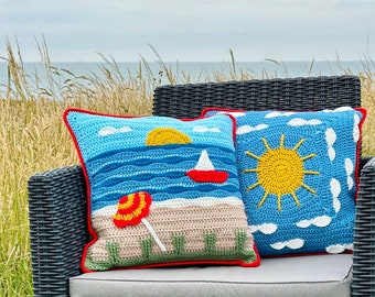 May include: Two crocheted throw pillows with summer themes. One pillow features a blue and white design with a yellow sun and white clouds. The other pillow is blue with a red sailboat and a yellow sun. Both pillows have red trim.