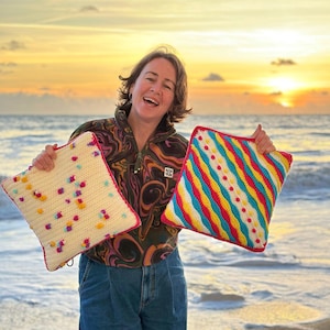 Op de afbeelding: Twee handgemaakte gehaakte kussens. Eén is crèmekleurig met kleurrijke bolletjes en een roze rand. De andere heeft diagonale strepen in rood, geel, blauw en wit, met een roze rand. Beide zijn vierkant.